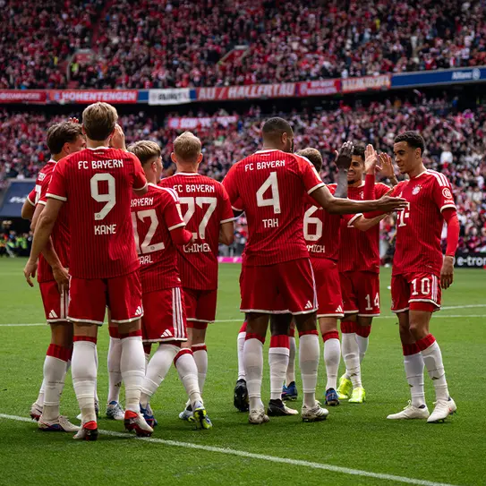 ფეხბურთელები FC Bayern-ის მაისურებით ერთმანეთს ჩახუტდებიან და ფართო მოედანზე მხარს უჭერენ გუნდის სულიერ აუარებას, ფონზე გადაღებული მრავალრიცხოვანი მაყურებელი.